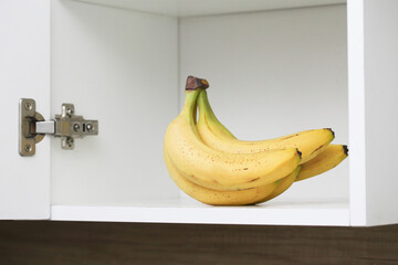 Bananas in kitchen cabinet. Yellow fruits in kitchen. Open door kitchen cabinet with hidden bananas inside. Ripe bananas with small brown stains. White empty copy space. Bunch of bananas isolated.