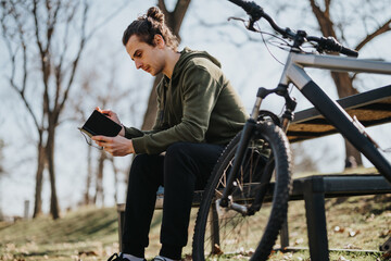 A good-looking young male relaxes on a park bench, reading with his bicycle beside him on a bright, sunny day.