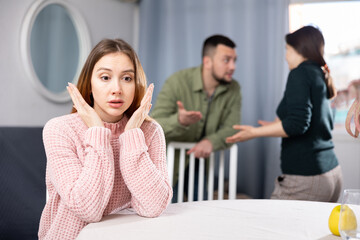 Stressed young woman sitting at table while family couple of her friends arguing behind - obrazy, fototapety, plakaty