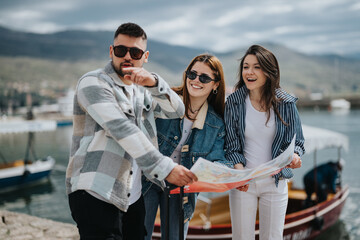 Three young adults enjoy a day out in a coastal town. A man guides two smiling women, using a map to explore the area.