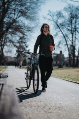 Casual young man stands with his bicycle in a park, holding a coffee cup, enjoying a beautiful sunny day outdoors.