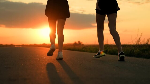 People run in sun together. Girls feet run along road training, , teamwork of running athletes. Slow motion. Athletic young women running along an asphalt road at sunset, healthy fitness lifestyle.