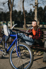 A bearded businessman enjoying a sunny day in the park, with a blue bicycle, working remotely on a tablet.