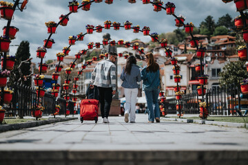 A young couple with a suitcase and their friend are walking under vibrant flower-filled arches in a scenic small town, enjoying a leisurely stroll and engaging in conversation.