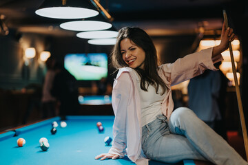Smiling young woman sitting by a pool table in a bar, radiating happiness and enjoying a playful night out with friends.