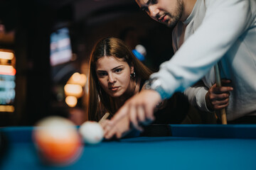 A focused woman takes aim during a friendly billiard game as a man assists with the cue, symbolizing leisure and togetherness.