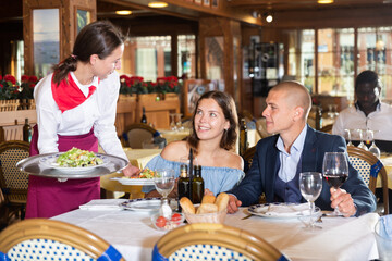 Smiling waitress serving food for positive couple at restaurant, putting plate with salad on table