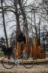 Young adult woman sitting on a unique wooden structure in a park, using a smart phone, with her bicycle nearby.