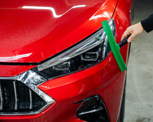 A man washes the headlights of a red car with a scraper. 