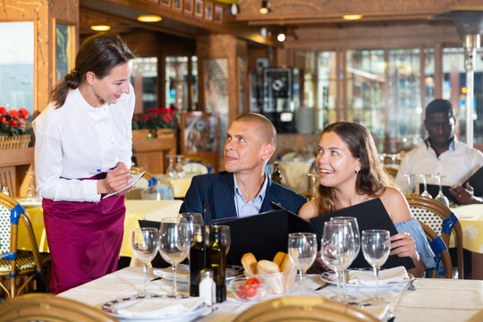 Attractive smiling woman waiter receiving order from guest in fashionable restaurant
