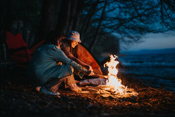 Two friends roast marshmallows over a cozy bonfire by the lake at dusk, showcasing a serene camping...