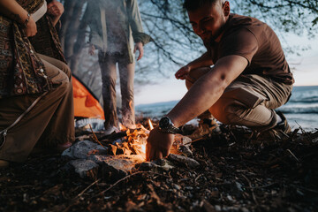 Group of friends enjoy a peaceful evening by the lake, warming their hands over a campfire as the sun sets.