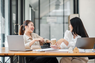 Two joyful businesswomen laughing and chatting over a cup of coffee at a wooden office desk with laptops.