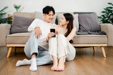 Young Asian couple sits cozily on a beige sofa, sharing a moment with a smartphone in a well-lit,...