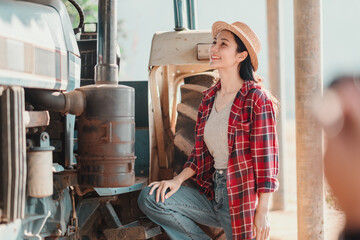 Joyful farmer in a straw hat and plaid shirt leans on a blue tractor, posing with hands on her hips. - Powered by Adobe