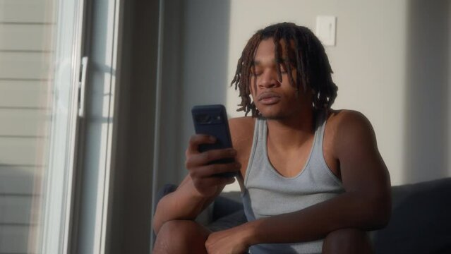 Young man with dreadlocks using his phone while sitting on a sofa next to the window. Slow motion. 