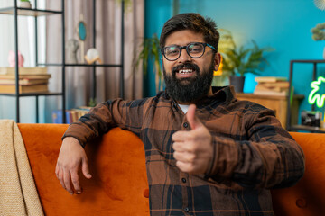 Like. Happy excited Indian man looking approvingly at camera showing thumbs up like sign positive something good great news positive feedback. Young Arabian guy sitting on couch at home living room