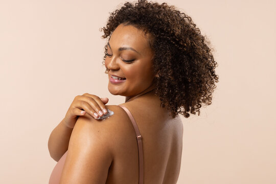 Biracial young female plus size model rubs lotion on shoulder, on beige background, copy space