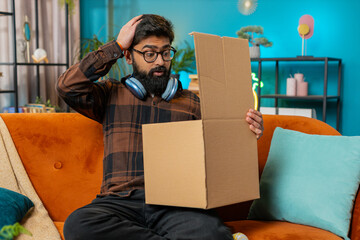 Angry dissatisfied shopper Indian man unpacking parcel feeling upset and confused with wrong mistake delivery from an online store, bad quality broken purchase at home. Guy indoors in room on couch