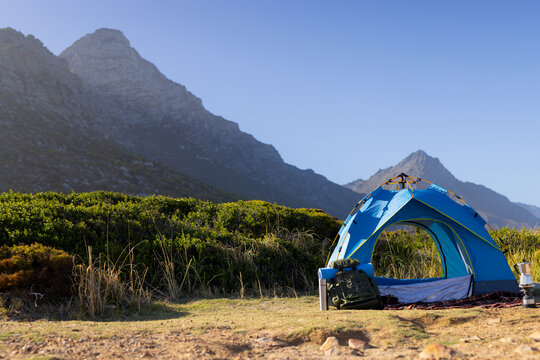 A blue tent sits at the foot of tall mountains, surrounded by greenery, copy space