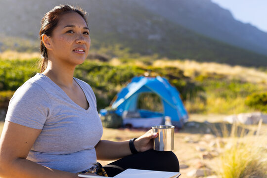 Biracial female hiker holding cup, sitting near a tent, copy space