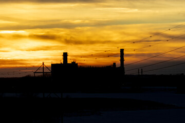 Sunsetting over industrial facility. Silhouette of large refinery with tall exhaust towers.  Power lines reach to the factory. Burning orange sky makes the background