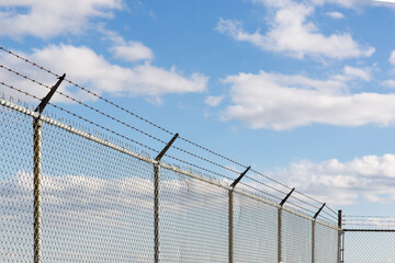 Barb wire on top of a chain link fence against a blue cloudy sky