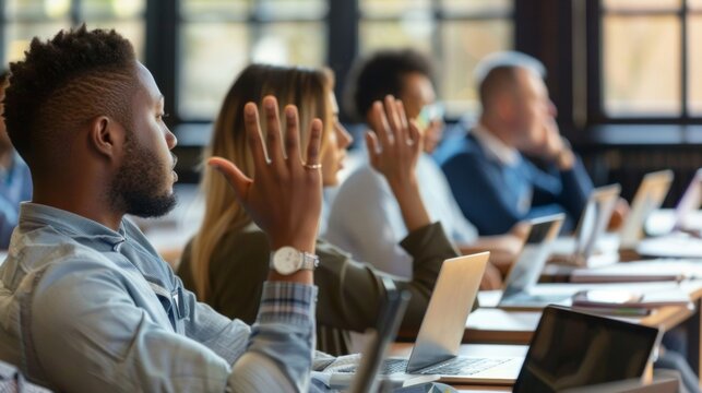 A group of employees sit in a conference room laptops open in front of them as they follow along with an online training program. One employee raises their hand to ask a question while .