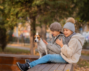 Brother and sister sit in an embrace with a dog on a bench for a walk in the autumn park. Boy, girl...