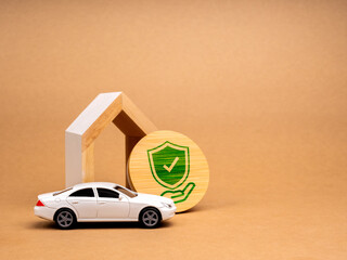 Home and car insurance concept. Hand holde green shield on wood badge with white toy car and modern house model is oled on brown background with copy space. Insurance or loan real estate or property.