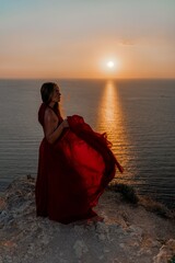 Woman sunset sea red dress, back view a happy beautiful sensual woman in a red long dress posing on a rock high above the sea on sunset.