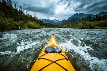  A person in a yellow kayak on the river faces the camera, with a dark green forest and mountains in the background. © Duka Mer