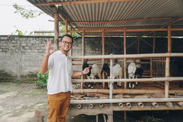 Excited young Asian man standing in front of traditional cage made from wood and bamboo in Indonesia rural area with goat inside