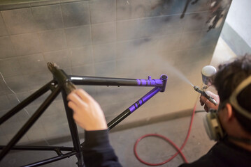 Man spray painting a bicycle frame in his workshop, applying a protective clear coat to give a...