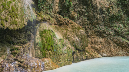 Fototapeta premium A beautiful tropical waterfall. Thin streams of water flow down the mossy stone terraces of the slopes, forming a veil. Turquoise lake at the foot of the cliff. Philippines. Cebu. Tumalog Falls.