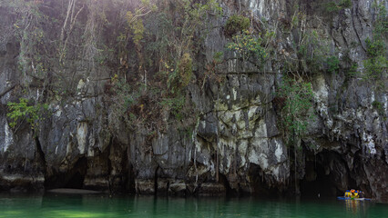 A canoe with tourists floats into the cave along the river. Sheer cliffs overhang the dark...