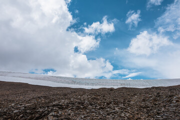 Minimalist layered landscape of stone hill with wide glacier in top against cirrus clouds in blue sky. Minimal nature backdrop with rock pass with ice cornice under cloudy sky in changeable weather.