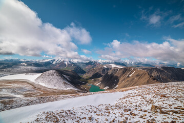 High snowy stony pass with vast view to most beautiful alpine lake, rocky hill in freshly fallen snow in sunlight and large colorful mountain range with snow-white peaks in far away in low clouds.
