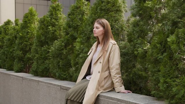 Three quarters view of a young woman in a light coat sitting on a parapet by evergreen trees and then jumping off.