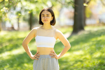 A woman is standing in a park, wearing a white tank top and grey sweatpants