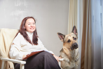 Middle aged Woman Relaxing in Chair With German Shepherd Indoors. Smiling mature woman in armchair with her attentive pet dog in cozy room