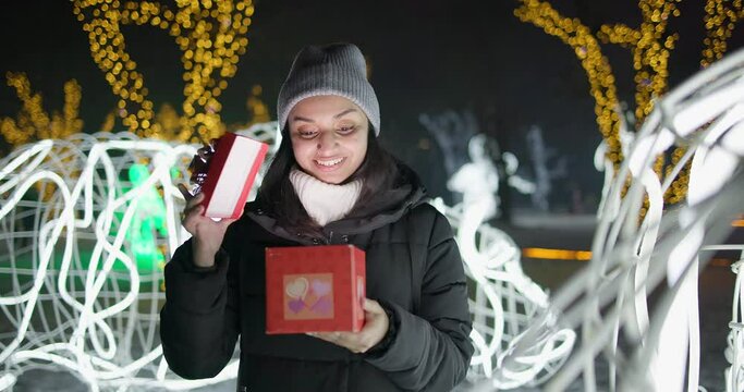 Young Indian woman opens gift box with bow standing on garlands and lights background outdoors - New Year holidays Christmas vacation