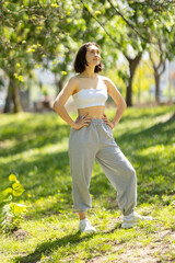 A woman is standing in a park, wearing a white tank top and grey sweatpants