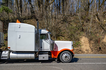 Extended cab white big rig bonnet semi truck with orange accents transporting wood boxes on flat bed semi truck running on the highway road with forest on the side