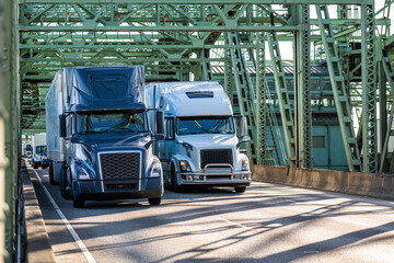 Two big rig semi trucks blue and white transporting cargo in dry van semi trailers running side by side on the truss arched bridge - obrazy, fototapety, plakaty