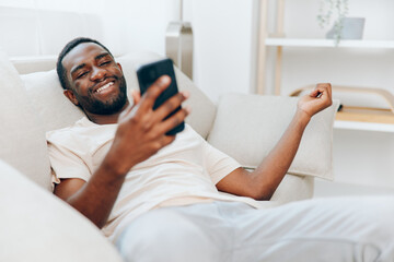 Happy African American Man Typing Message on Smartphone while Relaxing on Black Sofa at Home The modern apartment provides a cozy backdrop as the young man enjoys leisure time, confidently connecting