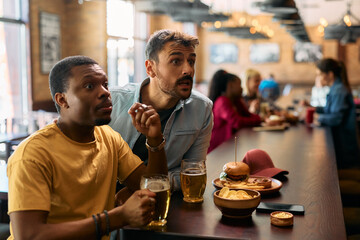 Young man and his African American friend watching sports game in  bar.