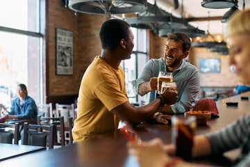 Happy man and his black friend toasting while drinking beer at bar counter.