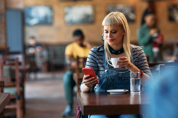 Fototapeta premium Smiling woman texting on cell phone while drinking coffee in cafe.
