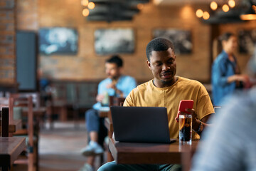 Young black man using mobile phone while surfing net on laptop in  cafe.